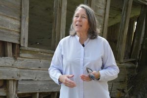A woman stands in the center of the frame with a weathered structure behind her, looking to her right and gesturing with her hands as she speaks.