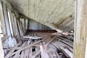 he interior of a deteriorating wooden structure. Floorboards are loose and scattered throughout the room. The ceiling has fallen, angling downward toward the right side of the room.