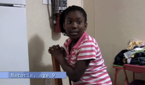 A 9-year-old girl in a pink-and-white striped shirt sits on a wooden chair.