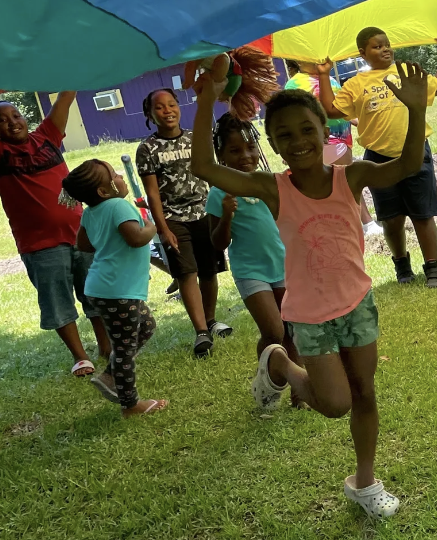 Children run and play under a colorful parachute.
