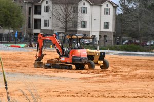 Two construction vehicles at rest on a cleared plot are centered in the frame. An apartment building is in the background.