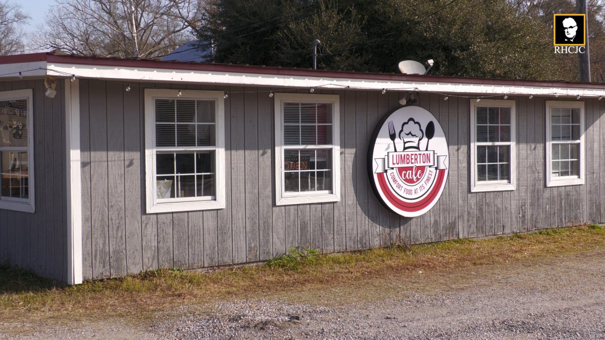 Exterior of Lumberton Cafe, a gray building with white trim and a round red-and-white sign mounted on the wall.