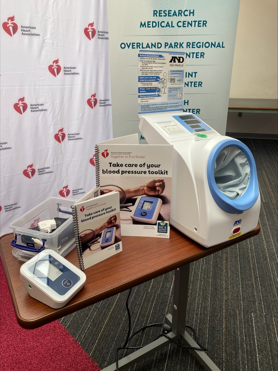 A table displays a public blood pressure monitoring machine, a digital cuff, and educational materials from the American Heart Association promoting blood pressure awareness.