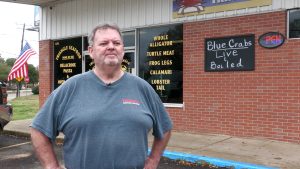 Man standing outside Triangle Seafood & Po’ Boy in front of a brick storefront and a sign advertising live and boiled blue crabs.