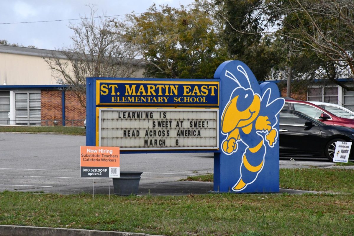 Sign for St. Martin East Elementary School with a hornet mascot and a marquee announcing, “Reach Across America March 6,” located in front of the school building and parking lot.
