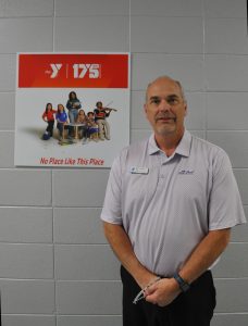 A man in a purple and white shirt and black pants stands in front of a painted cinder block wall with a red-and-white sign marking the Y's 175th anniversary.