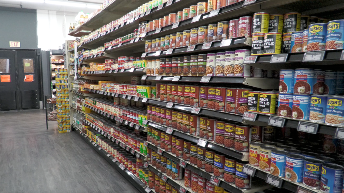 Shelves in a grocery store aisle are stocked with canned beans, vegetables and other foods, with price tags posted along the shelves.
