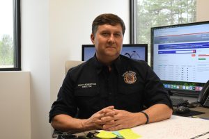 Trent Robertson sits at a desk and smiles at the camera. His shirt reads “Trent Robertson, Director” and displays a Greene County Emergency Management logo. Two computer monitors are behind him.