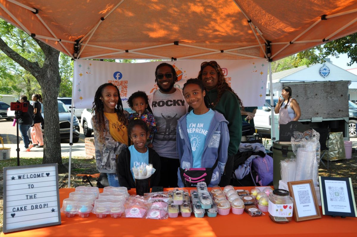 A family stands behind a table of baked goods under an orange tent with a sign that reads, “Welcome to the Cake Drop!”