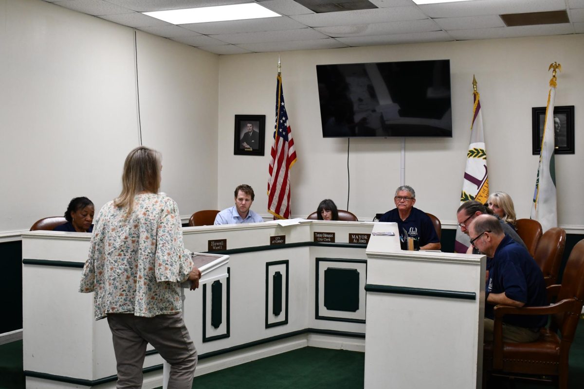 Members of the Lucedale Board of Aldermen sit at the front of a meeting room as a woman speaks at a podium.