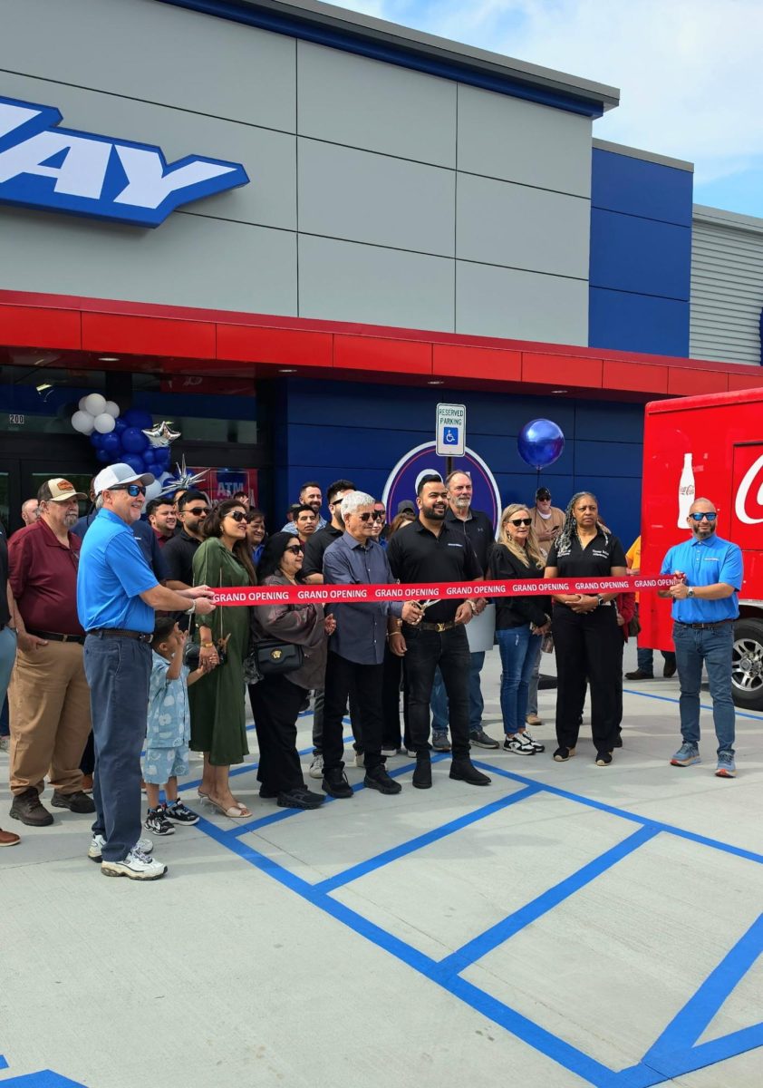 A crowd stands outside a gas station holding a red ribbon during a grand opening ceremony.