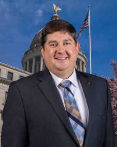 Sen. Jeremy England poses for a headshot in a suit and ties with the Mississippi Capitol in the background.