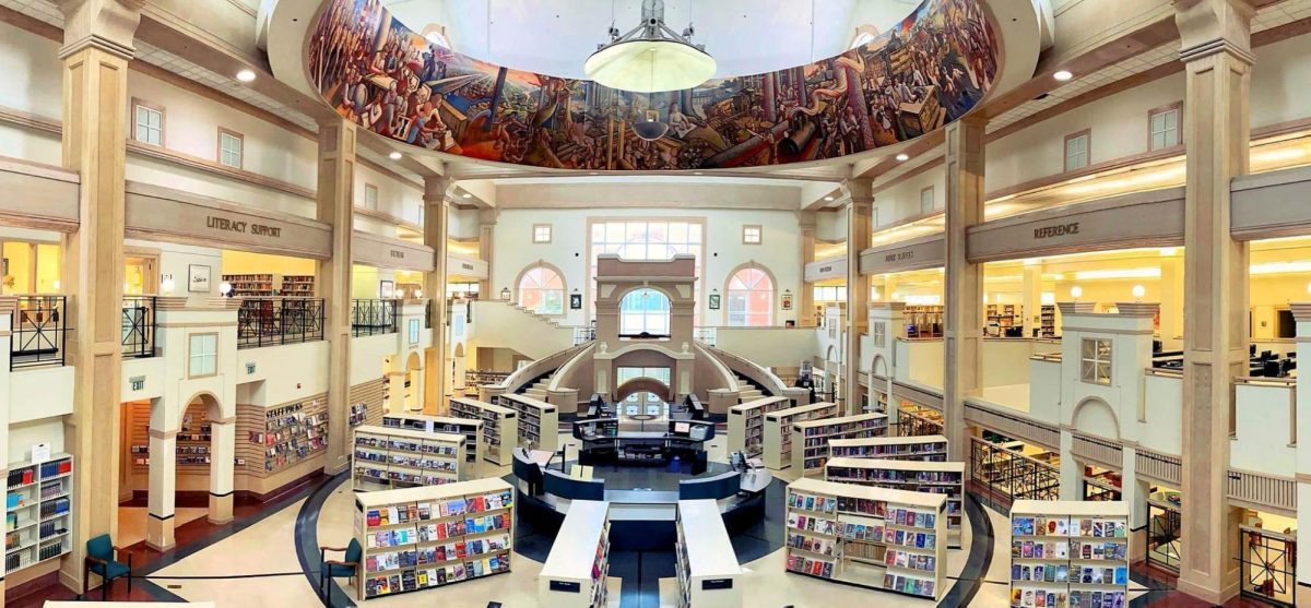 Interior view of the Library of Hattiesburg, Petal and Forrest County showing book stacks, upper-level reading areas, a central service desk, sweeping staircases and a large circular mural above the main floor.