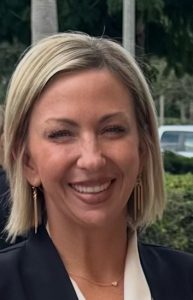 ALT Text: A blonde woman with short hair wears a black jacket and white shirt and stands in front of a bush, smiling at the camera.