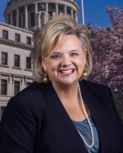 A woman wearing a black blazer and pearls smiles for a headshot with the Mississippi Capitol as the backdrop.