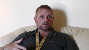 A bearded man in a dark polo sits on a couch talking to someone out of frame.