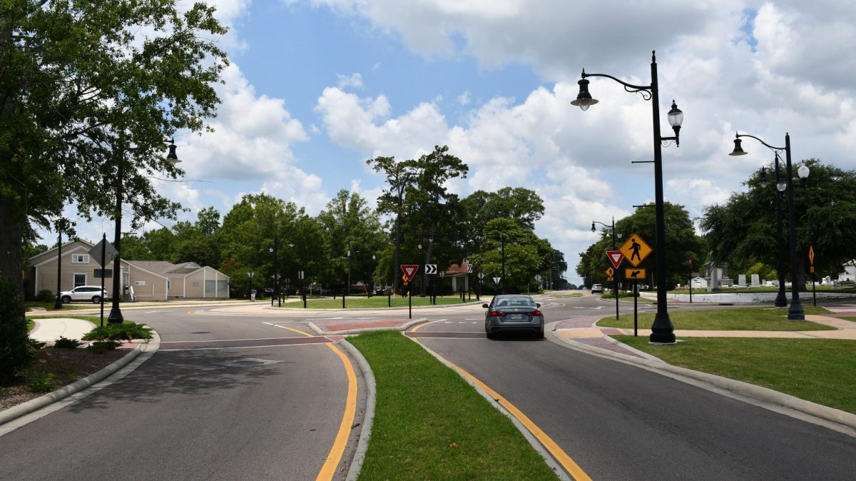 A car travels through a landscaped roundabout on a wide paved road lined with black streetlights, traffic signs, trees and a few buildings under a partly cloudy sky.