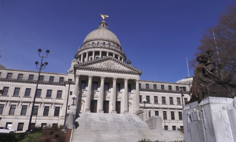 The Mississippi State Capitol building in Jackson, with a domed roof, tall columns and front steps under a clear blue sky.