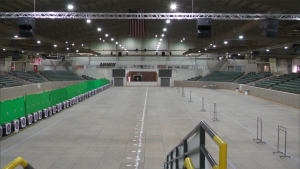 A large indoor arena with stadium seating and archery targets along one side.