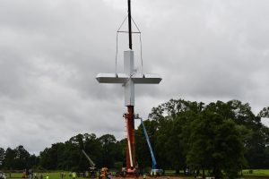 A crane lowers the top section of a large white cross onto its base along U.S. Highway 98 near the George-Greene county line.