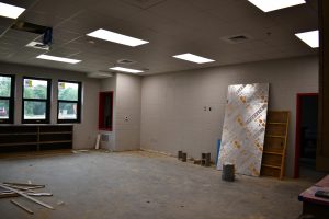 An unfinished classroom has bare floors, ceiling lights, windows and construction materials inside.