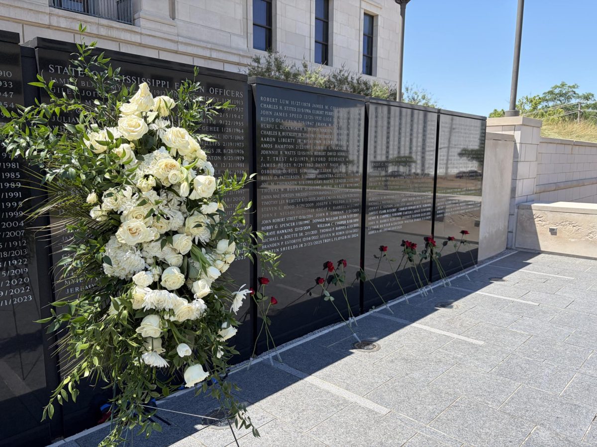 A white floral wreath stands in front of the Mississippi Fallen Officers Memorial Wall, where red roses read against the black stone panels.