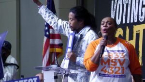 A woman speaks into a microphone in front of a podium as a man raises his arm with his eyes closed.