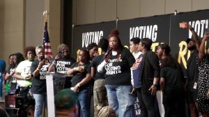 A woman with a microphone stands in front of a group of people, with onlookers and a video camera in the foreground.