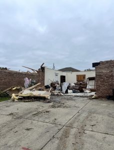 A storm-damaged brick home with part of its roof and walls missing. Broken wood, insulation, furniture and other debris are scattered across a driveway under a cloudy sky.