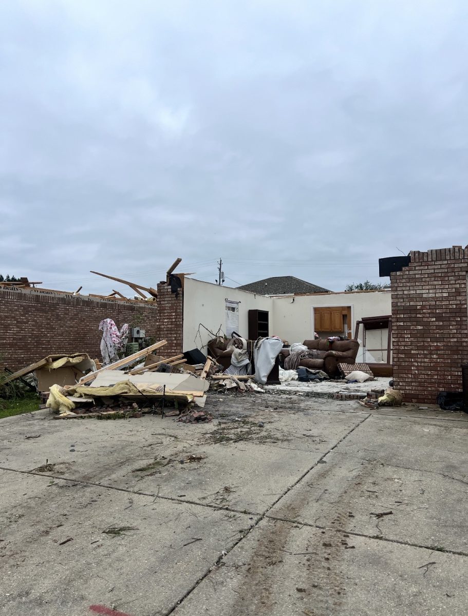 A storm-damaged brick home with part of its roof and walls missing. Broken wood, insulation, furniture and other debris are scattered across a driveway under a cloudy sky.