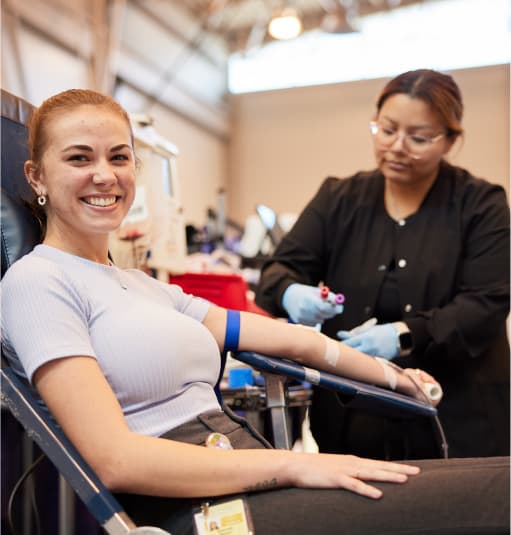 A smiling blood donor sits in a reclining chair while a health care worker prepares donation supplies nearby.