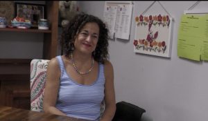 A woman sits at a desk in an office, wearing a blue-and-white striped sleeveless top. Behind her are shelves and papers posted on a wall.