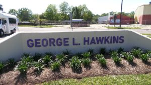 A white monument sign with purple letters spelling “George L. Hawkins” stands in a landscaped area outside a school campus.