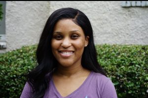 A woman wearing a purple shirt stands in front of a tan and green background.
