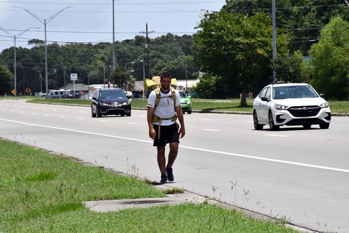 A backpacker walks beside a busy highway on a sunny day with cars passing in adjacent lanes.