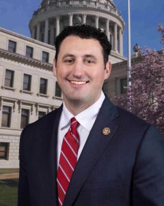 Rep. Jimmy Fondren smiles in a formal portrait while wearing a dark suit and red striped tie, with the Mississippi Capitol in the background.