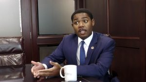 Rep. Justis Gibbs, wearing a blue suit and tie, gestures while speaking during an interview in a conference room.