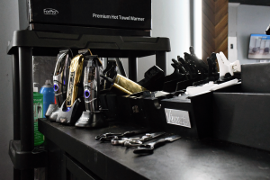 A black barber workstation holds several electric clippers, clipper guards, combs and shears, with a hot towel warmer above it.