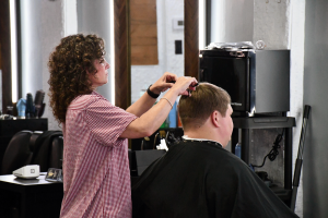 Crystal McCormick, wearing a red-and-white checkered shirt, trims the top of a seated client’s hair inside a barbershop.