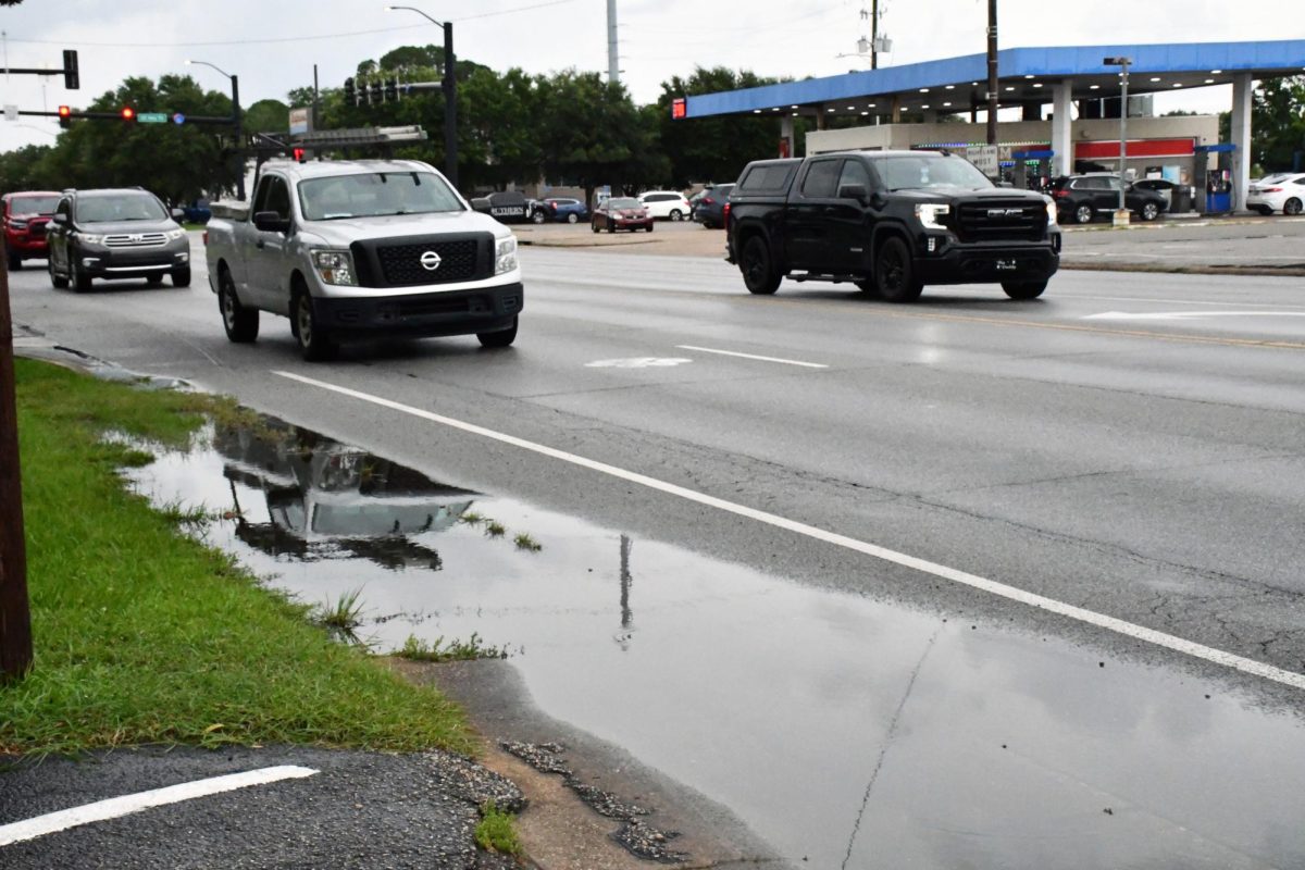 Several vehicles travel along a wet, multi-lane roadway while a large puddle sits beside the curb next to a grassy shoulder.