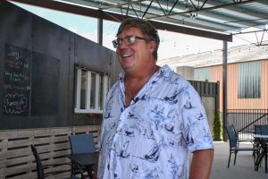 A man wearing glasses and a patterned button-down shirt stands smiling beneath a covered outdoor patio area.