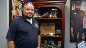 A man in a dark police polo shirt stands beside a wooden display case filled with framed photos, police patches, badges, a cap and other memorabilia.