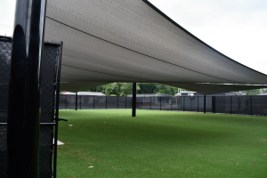 A fenced outdoor area with green turf is covered by large black shade sails attached to tall poles.
