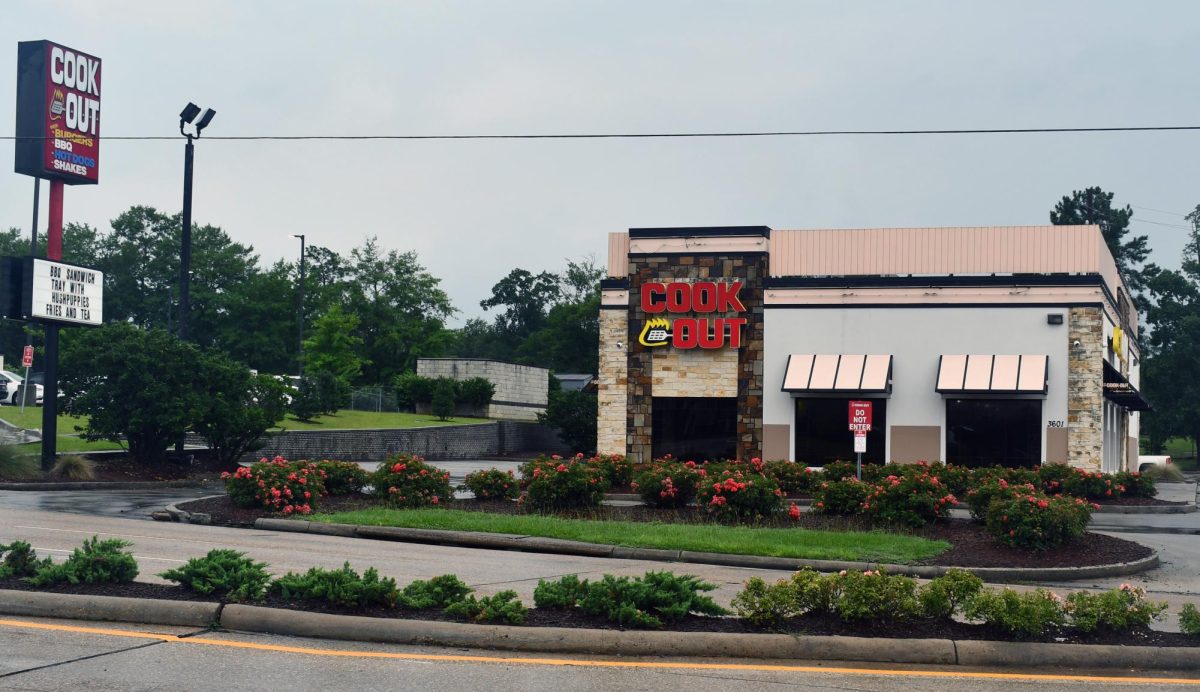 A Cook Out restaurant sits along Hardy Street in Hattiesburg, Mississippi, on a cloudy day. The building features the chain’s red-and-yellow sign above the entrance, with landscaped flower beds and a roadside Cook Out sign visible in front of the restaurant.