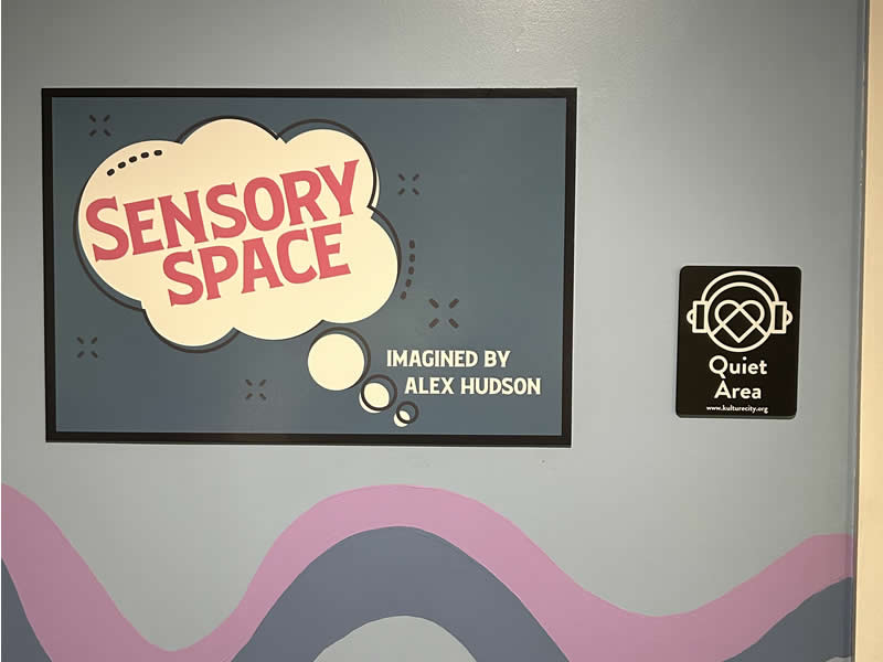 A wall at Lynn Meadows Discovery Center features a sign reading “Sensory Space” with a thought bubble design and a “Quiet Area” sign nearby.