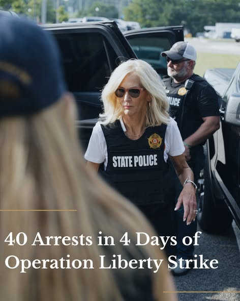 Lynn Fitch in focus wearing a police vest reading “STATE POLICE” and a badge in front of a car and male police officer with a long-haired officer standing in a cap facing away from the camera taking up the bottom left of the image.