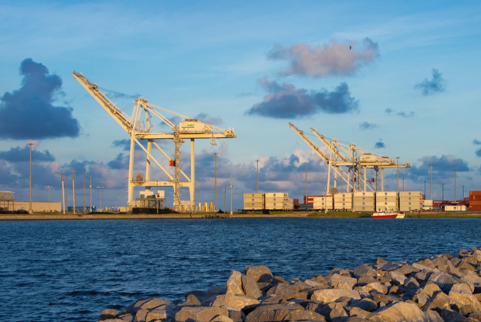 Container cranes, stacked shipping containers and cargo facilities at the Port of Gulfport are seen across the water, with a rocky shoreline in the foreground.