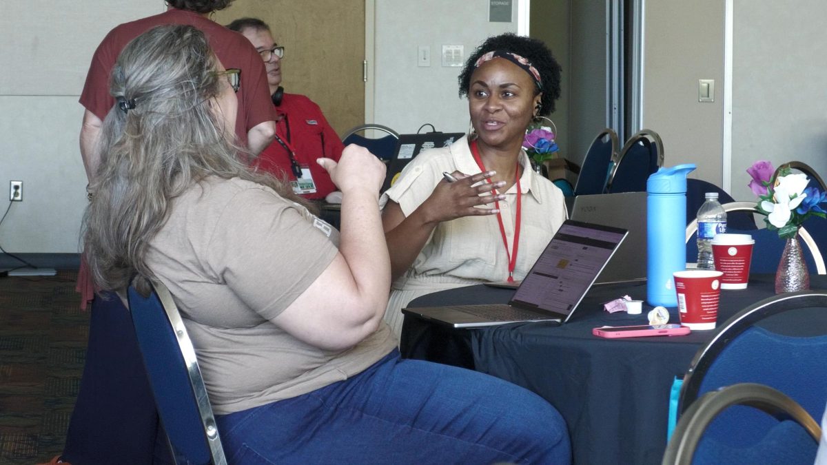Two women sit at a round table and talk while working on laptops during a conference. Other attendees are visible in the background.