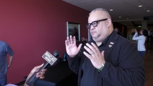 A man wearing glasses and a dark jacket gestures with both hands while speaking during an interview. A microphone is held toward him.
