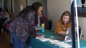 One woman leans over a table covered with brochures while another woman sits behind a laptop and points to information on the screen.